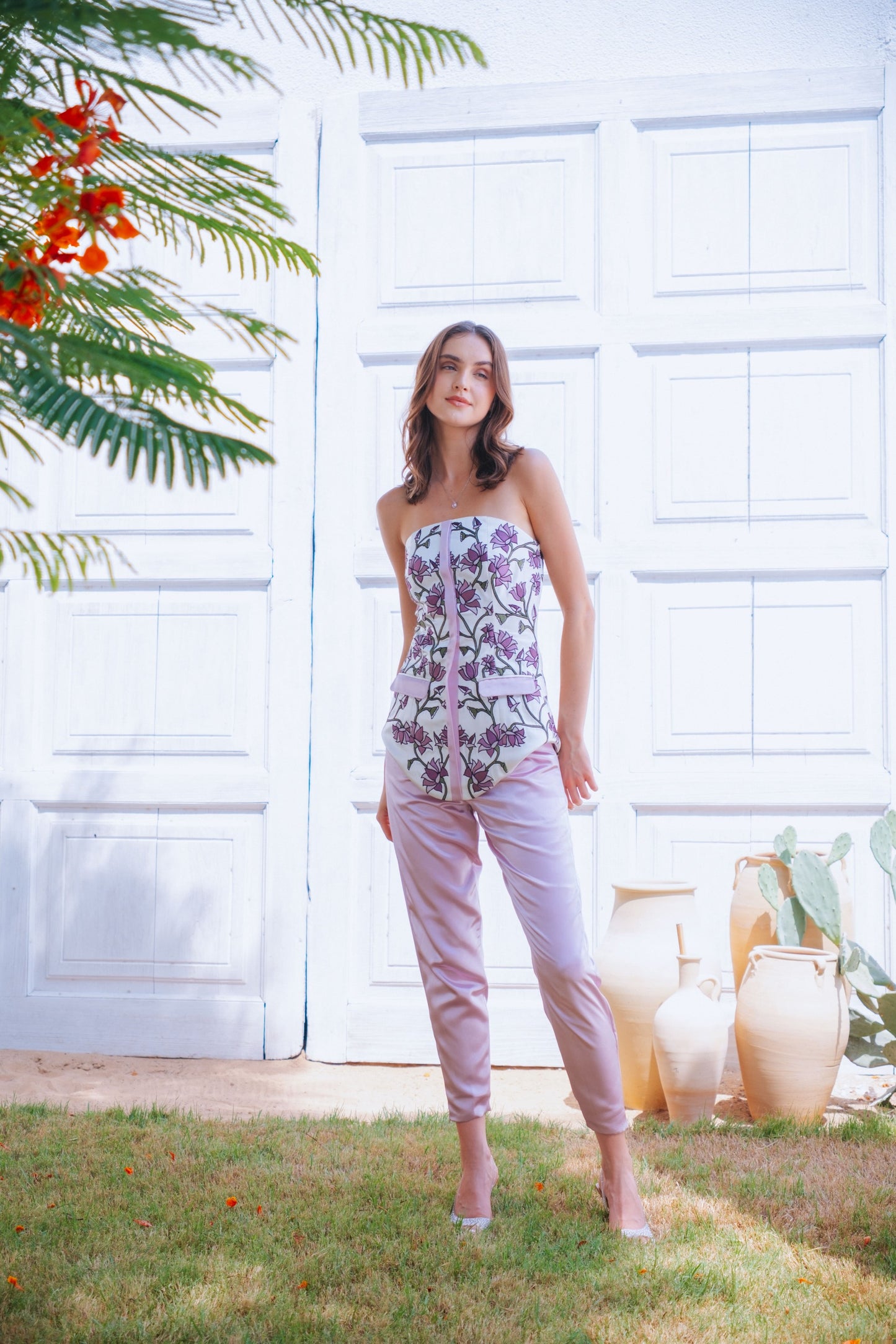Woman in a floral top and light pink pants standing outdoors with a white wall and greenery in the background