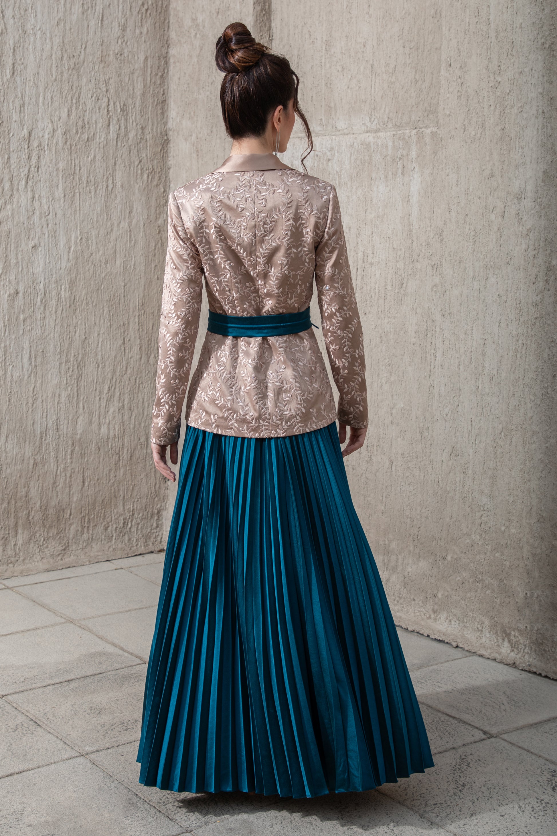 back -pose -showing -blue -pleated skirt-an -embroidered- brown-top