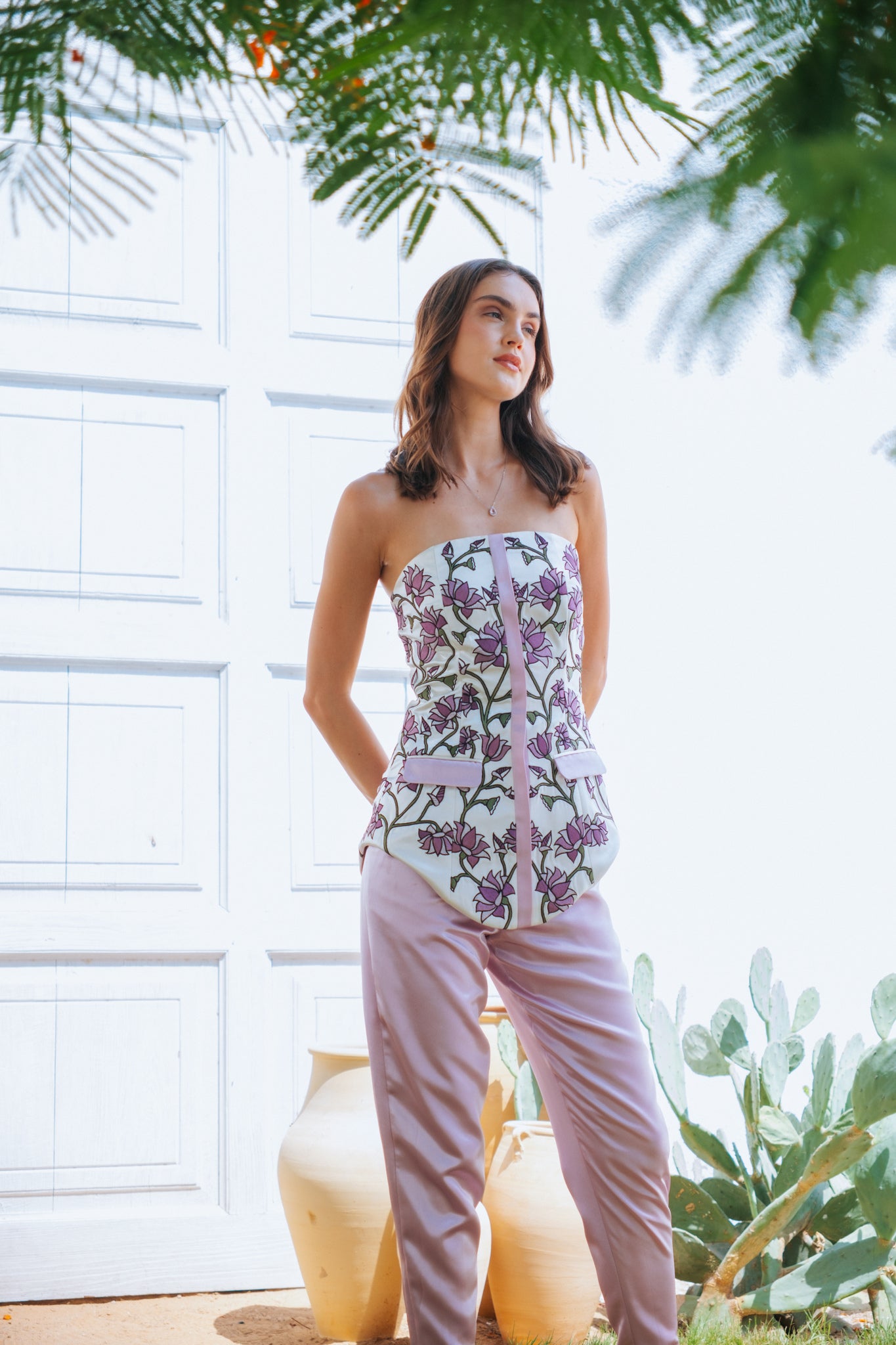 Woman wearing a floral strapless top and pink pants standing outdoors with plants in the background