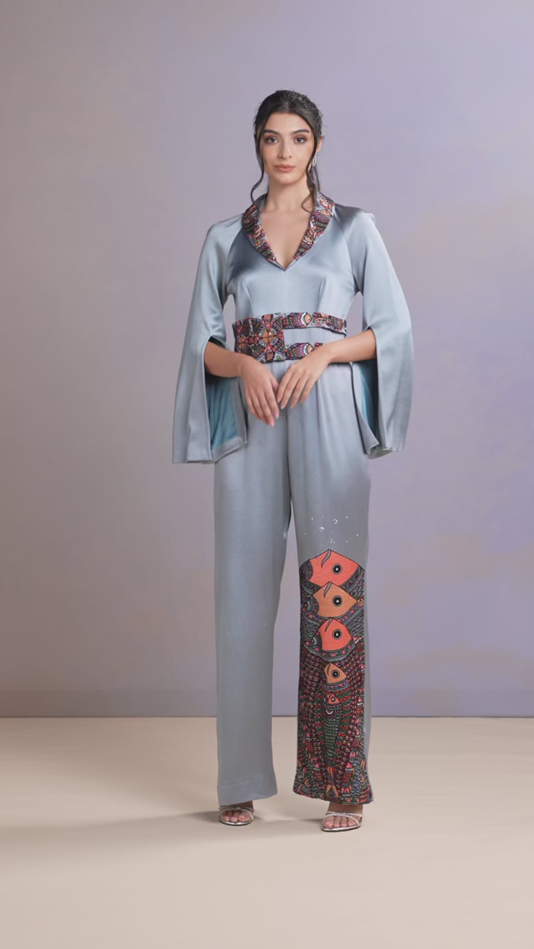 Model walking in glacier blue silk jumpsuit with floral embroidery and cape sleeves