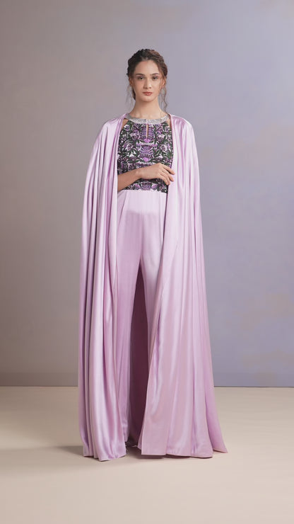 Model walking in French lilac satin jumpsuit with embroidered top and cape