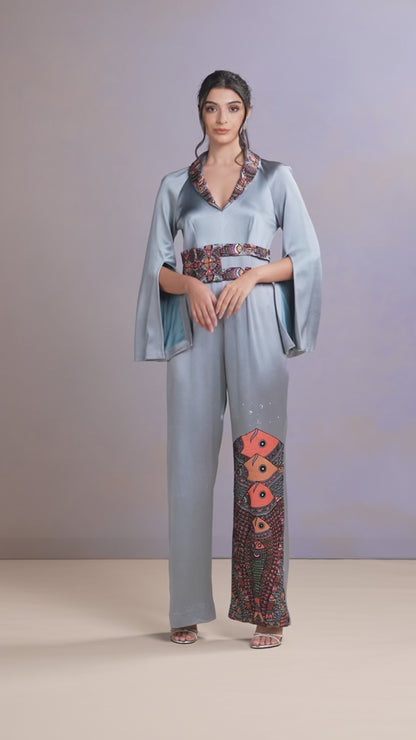 Model walking in glacier blue silk jumpsuit with floral embroidery and cape sleeves